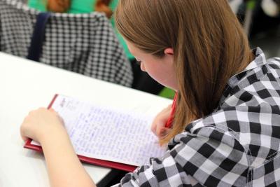 A photo of a girl, over he shoulder, looking down on a sheet of paper she is writing on. 