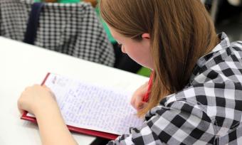 A photo of a girl, over he shoulder, looking down on a sheet of paper she is writing on. 